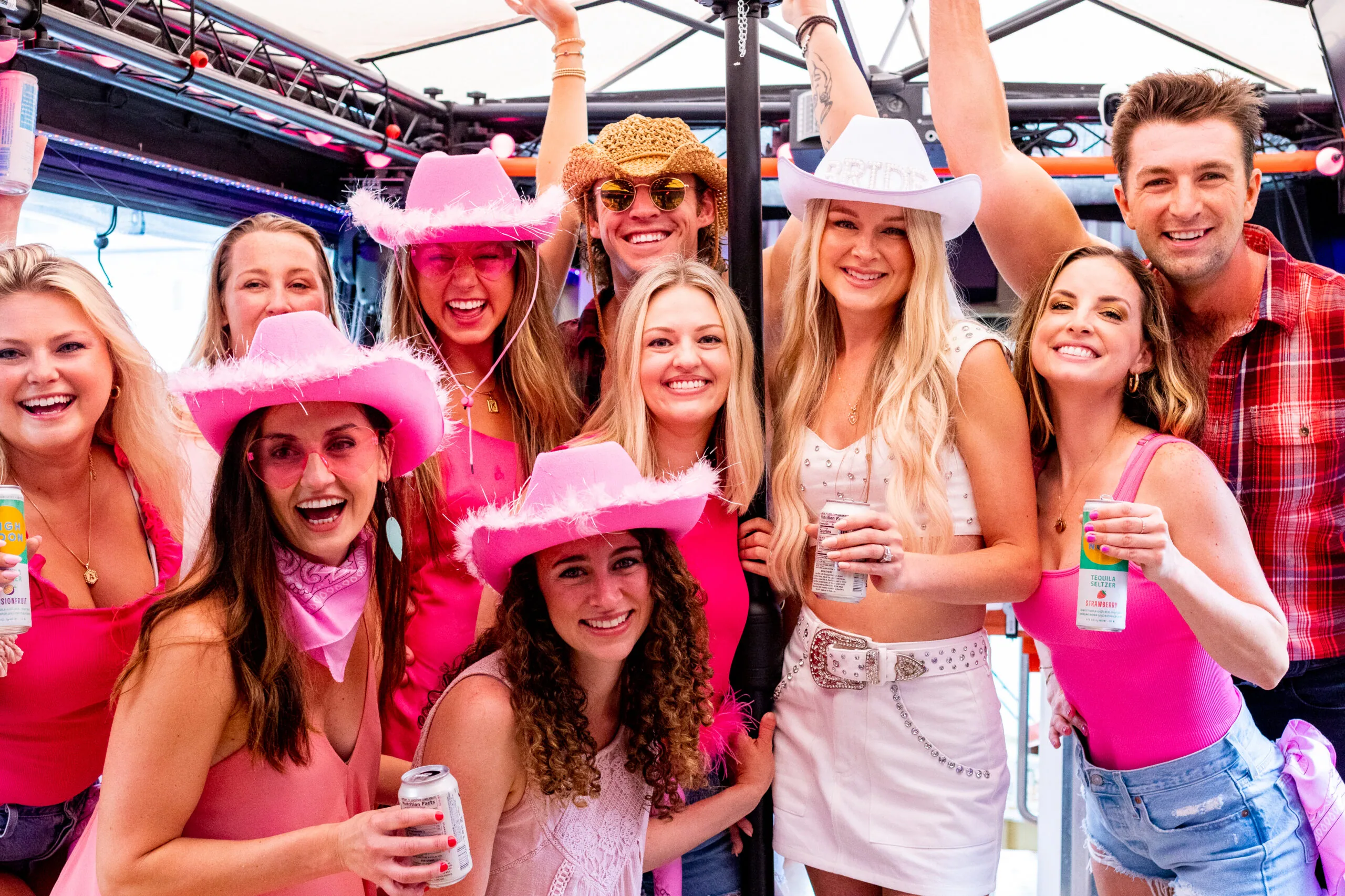 A lively bachelorette party group dressed in pink outfits and feathered cowboy hats cheers and smiles together under a covered outdoor venue, with the bride wearing a white rhinestone "Bride" cowboy hat and matching outfit, holding canned drinks.