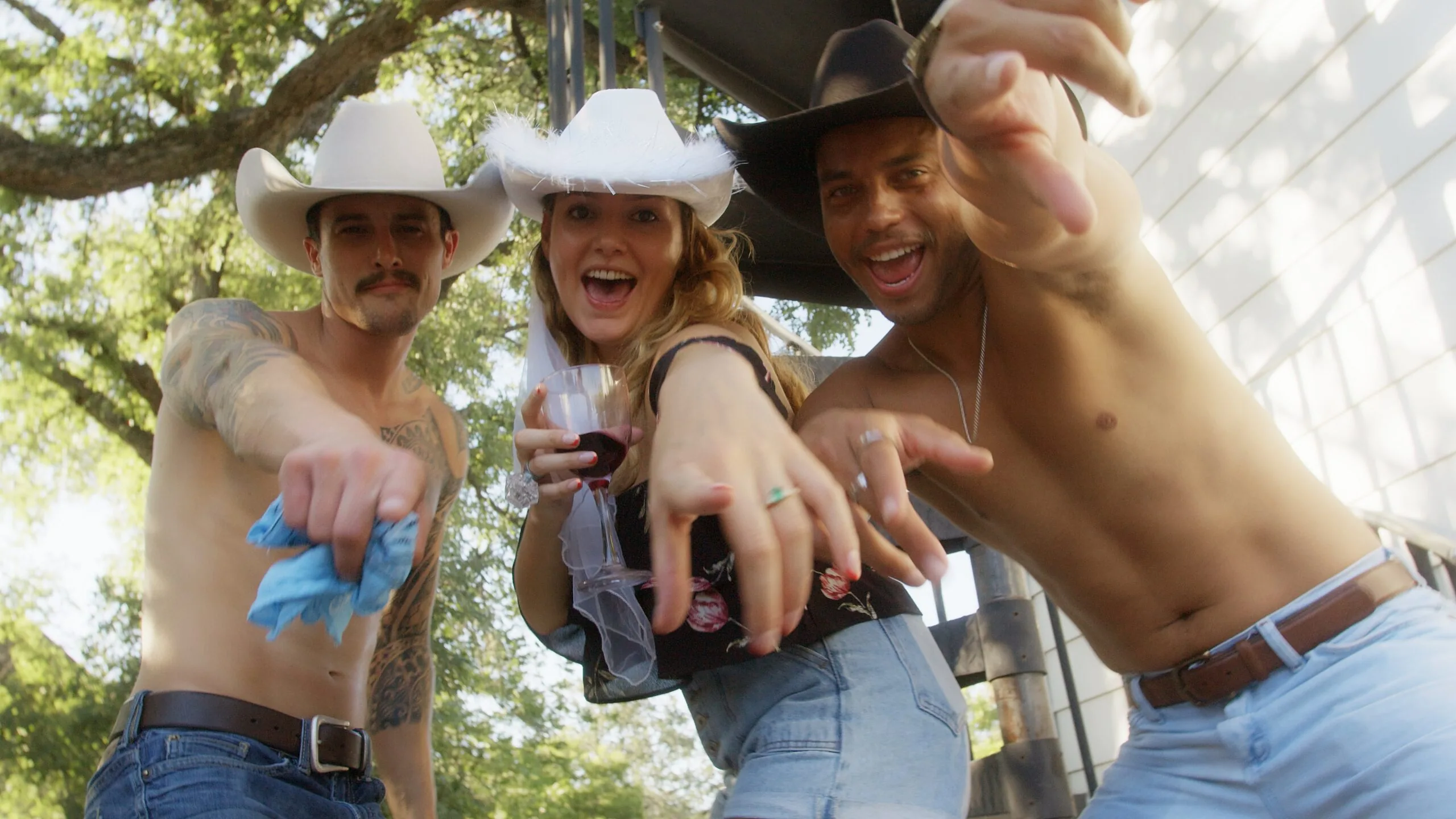 A bride-to-be in a white cowboy hat and veil points at the camera with two shirtless Cocktail Cowboys at a Nashville bachelorette party.