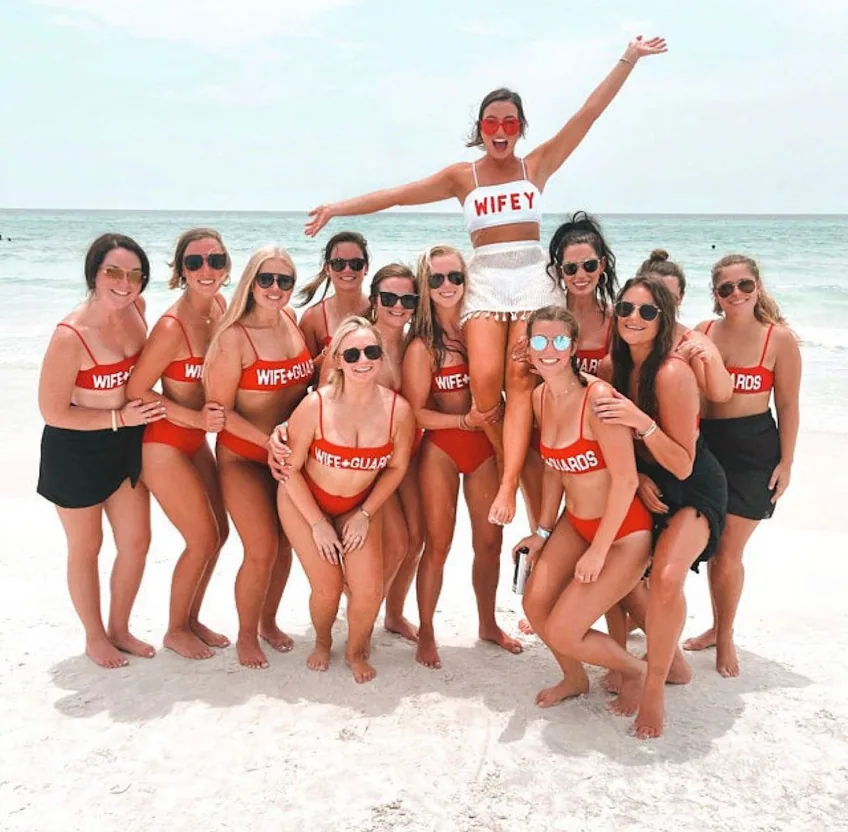 Large bachelorette group in swimwear posing on a sunny beach with arms raised