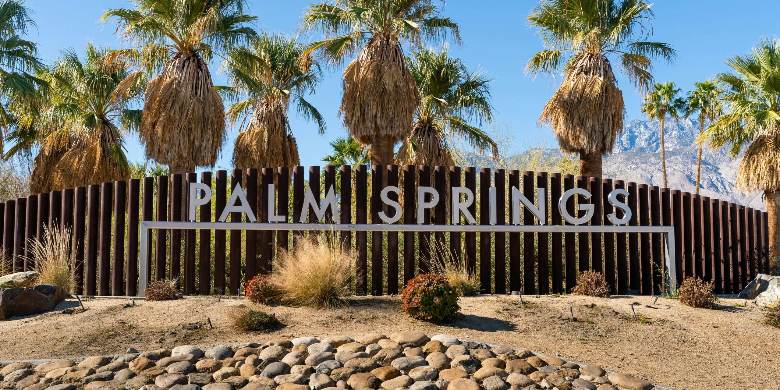 The Palm Springs welcome sign in silver letters mounted on a dark wooden slat fence, surrounded by desert landscaping, fan palm trees, and snow-dusted mountains under a clear blue sky.