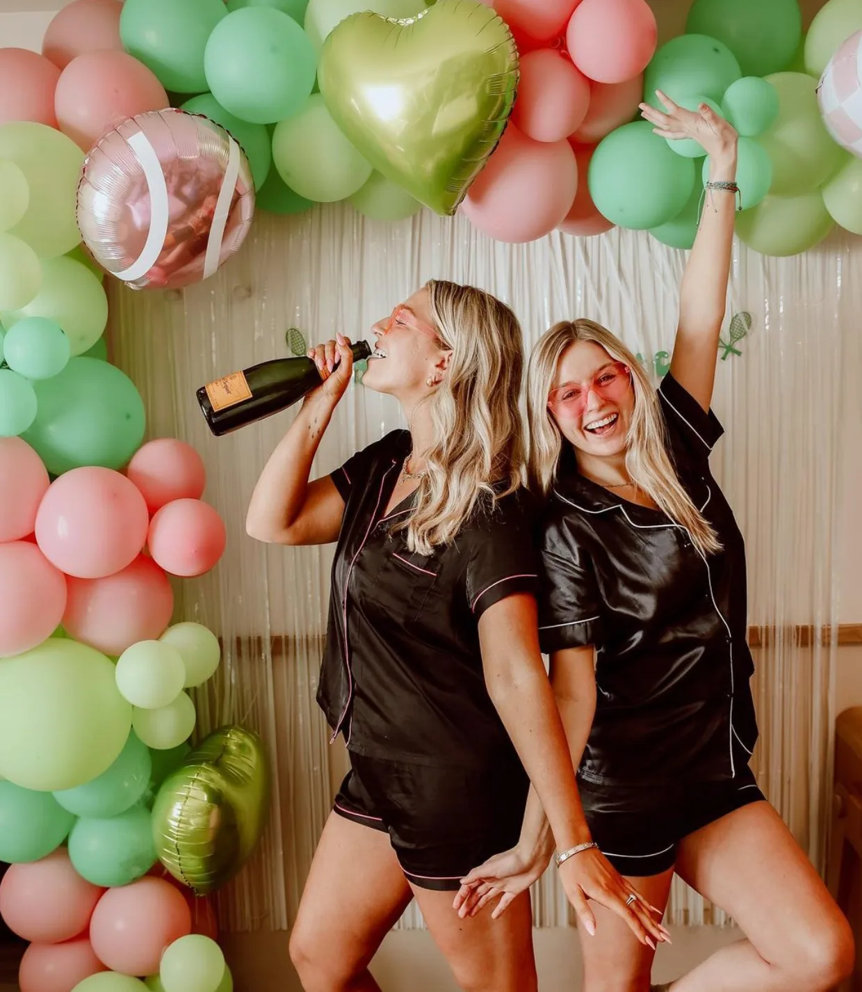Two women celebrating in front of a colorful green and pink balloon wall at a party