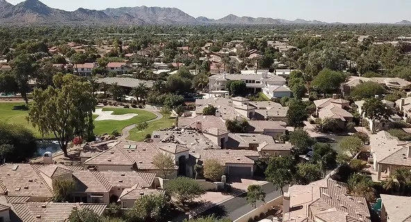Aerial daytime view of a Scottsdale residential neighborhood with tan tile-roofed homes, a golf course fairway with sand traps, mature trees, and the McDowell Mountains stretching across the horizon