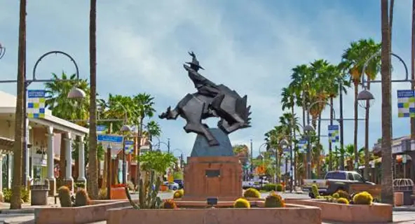 Bronze bucking bronco and cowboy sculpture centered on a stone pedestal along Old Town Scottsdale's palm tree-lined main street, with southwest-style shops and colorful Scottsdale city banners in the background
