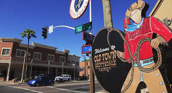 Welcome to Old Town Scottsdale sign featuring a painted cowboy with a lasso at the Main Street intersection, with southwest-style brick storefronts and palm trees under a clear blue sky