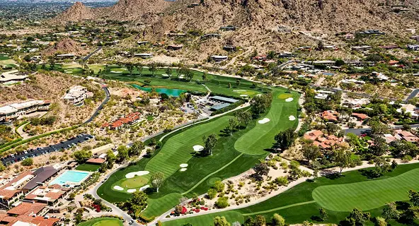 Aerial view of a manicured golf course with sand traps and a green pond winding through a luxury desert community at the base of rugged rocky mountains in Scottsdale, Arizona