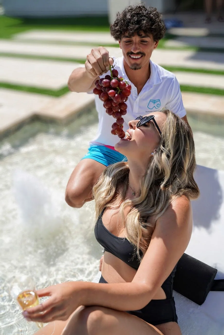 Cabana Boys host playfully popping champagne near a laughing female guest in a pool