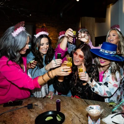 A bachelorette group in pink and blue outfits celebrating with drinks at an indoor party table