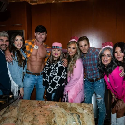 A large mixed group posing with Cocktail Cowboys hosts at a dimly lit indoor bachelorette party