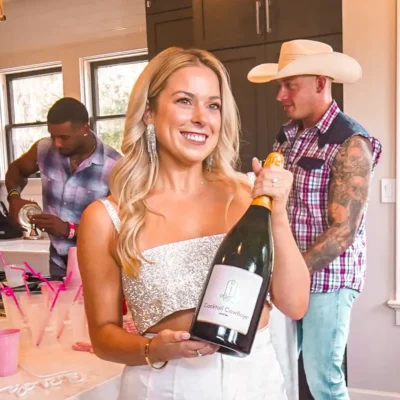 A smiling woman in a white sequin top holding a wine bottle next to a Cocktail Cowboys host in a straw hat