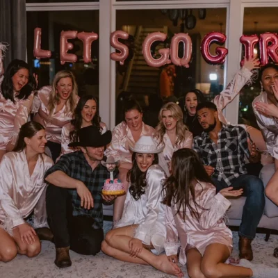 A bachelorette group in robes posing with Cocktail Cowboys hosts in front of a "Let's Go Girls" neon sign