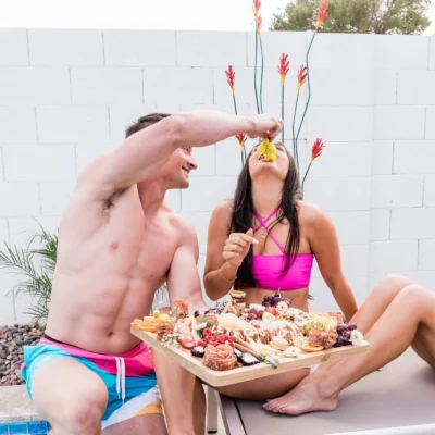 Shirtless Cabana Boys host presenting a charcuterie board to a seated female guest poolside