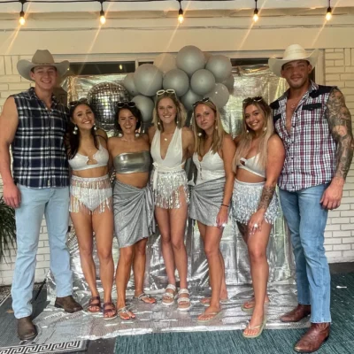 A large bachelorette party group in coordinating outfits posing with two Cocktail Cowboys hosts outdoors