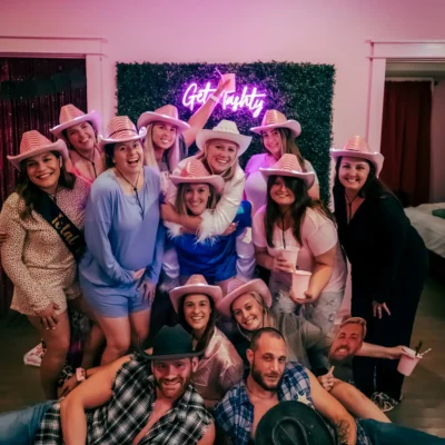 A large bachelorette group in pink hats posing with Cocktail Cowboys hosts in front of a neon "Girls" sign indoors