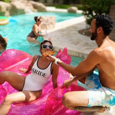 Cabana Boys host pouring champagne for a woman in a "Bride" sash floating on a pink pool float