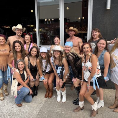 A large bachelorette group posing with Cocktail Cowboys hosts outside a venue entrance, all kneeling and smiling for a group photo