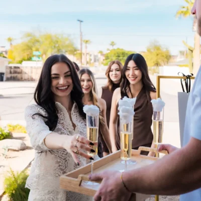 A Cabana Boys host handing champagne flutes to three women in coordinating brown outfits outdoors