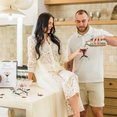 A Cocktail Boys host in a white shirt pouring wine for a woman in a white lace dress in a bright kitchen