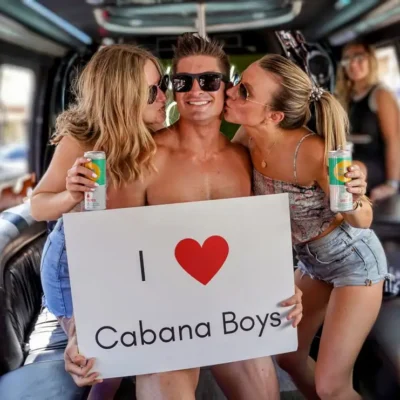 A shirtless Cabana Boys host in colorful shorts posing with an "I Love Cabana Boys" sign inside a party bus