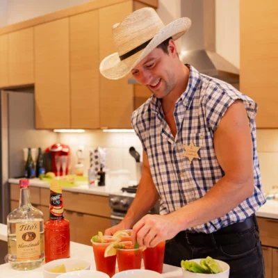 A Cocktail Cowboys host in a straw hat and plaid shirt preparing drinks at a kitchen counter with liquor bottles