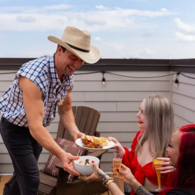 A Cocktail Cowboys host in a straw hat serving food to a woman in a red top at an outdoor table