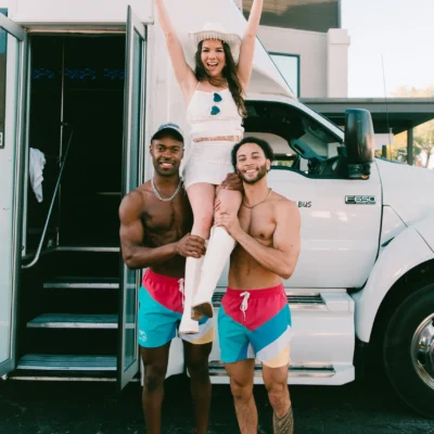 Two shirtless Cabana Boys hosts in colorful shorts posing in front of a white party bus
