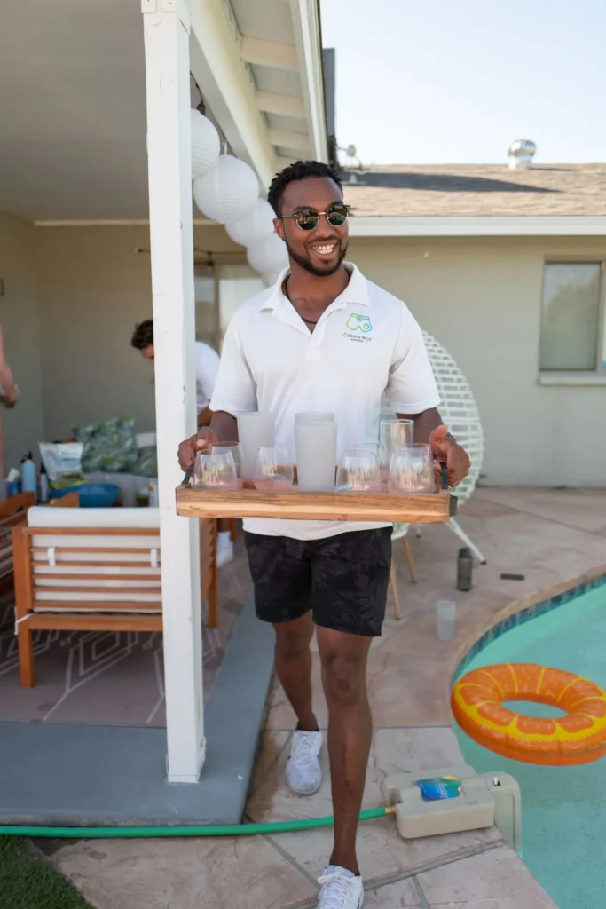 A Cabana Boys host in a white polo carrying a tray of shot glasses at an outdoor pool party