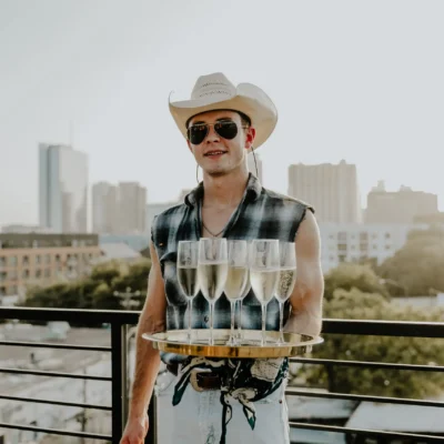 Two Cocktail Cowboys hosts in plaid shirts and cowboy hats holding champagne flutes on a Miami rooftop with city skyline views