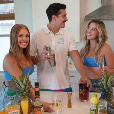 A Cabana Boys host in a white polo serving tropical drinks and a pineapple to two women outdoors