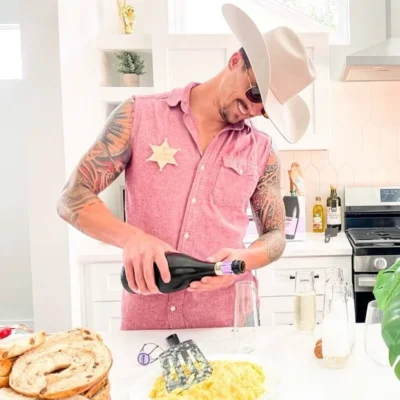 A Cocktail Cowboys host in a pink plaid shirt and sheriff star badge pouring champagne over a food spread in a bright kitchen