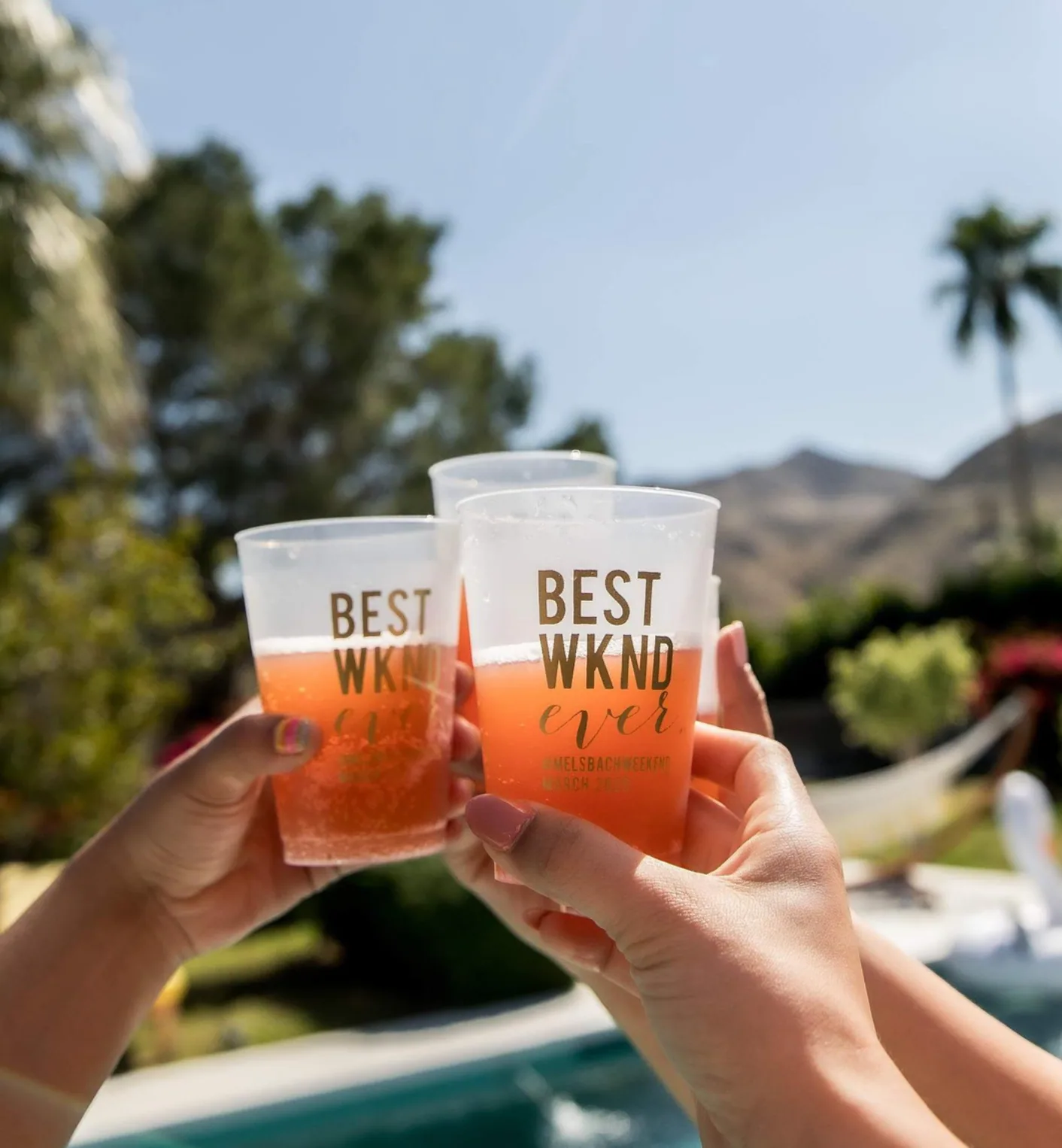 Two hands toasting with frosted plastic cups printed with "Best Wknd" against a bright outdoor background with palm trees