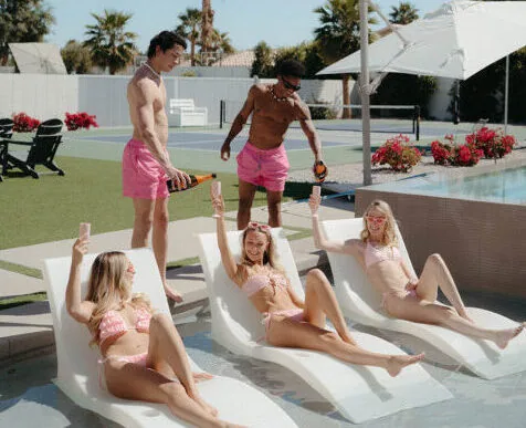 A Cabana Boys host in pink shorts pouring a drink for women relaxing on pool ledge chairs at a sunny outdoor pool