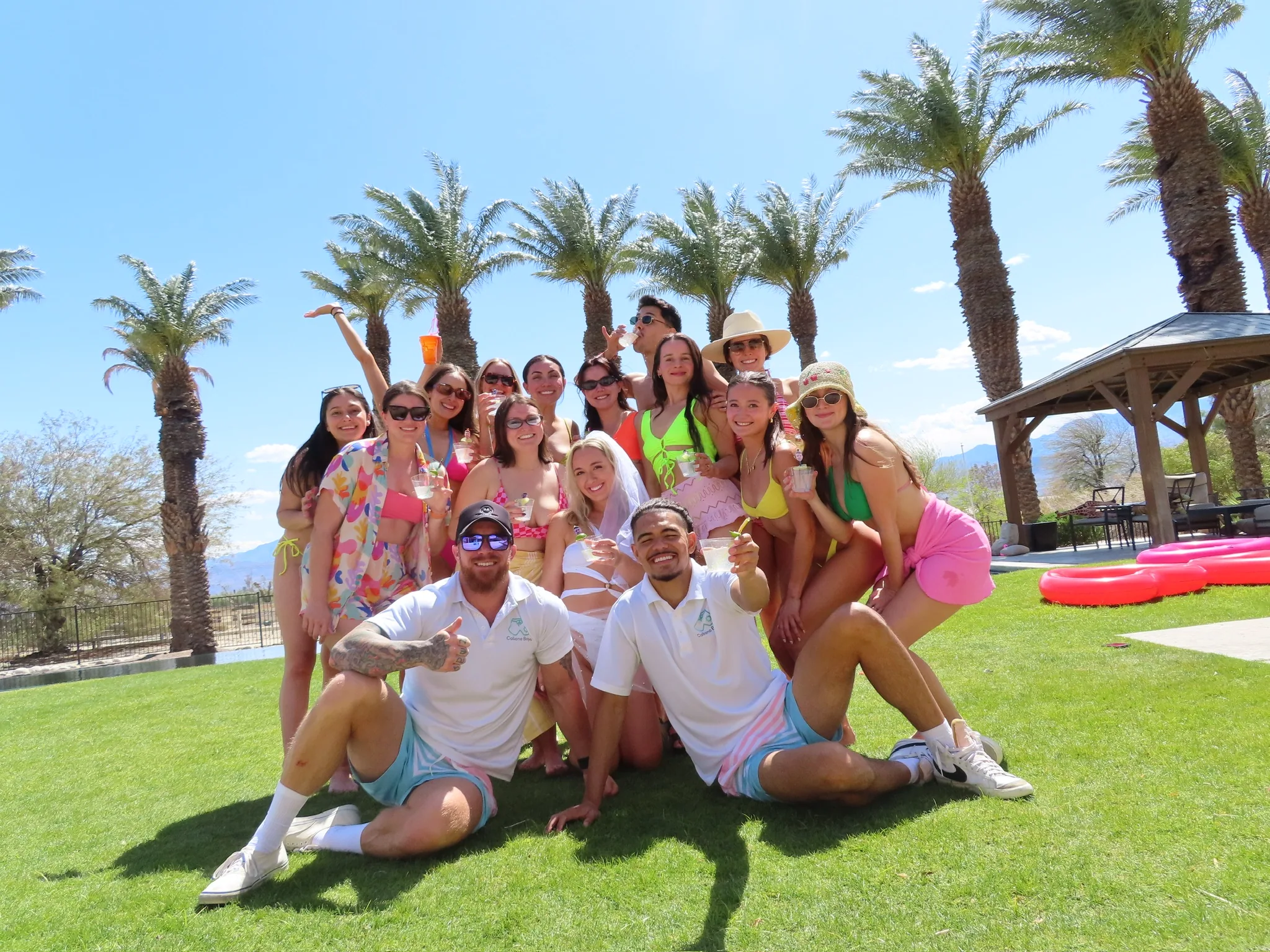 A large bachelorette party group posing with two Party Host Boys on a lawn surrounded by palm trees and bright pink pool floats