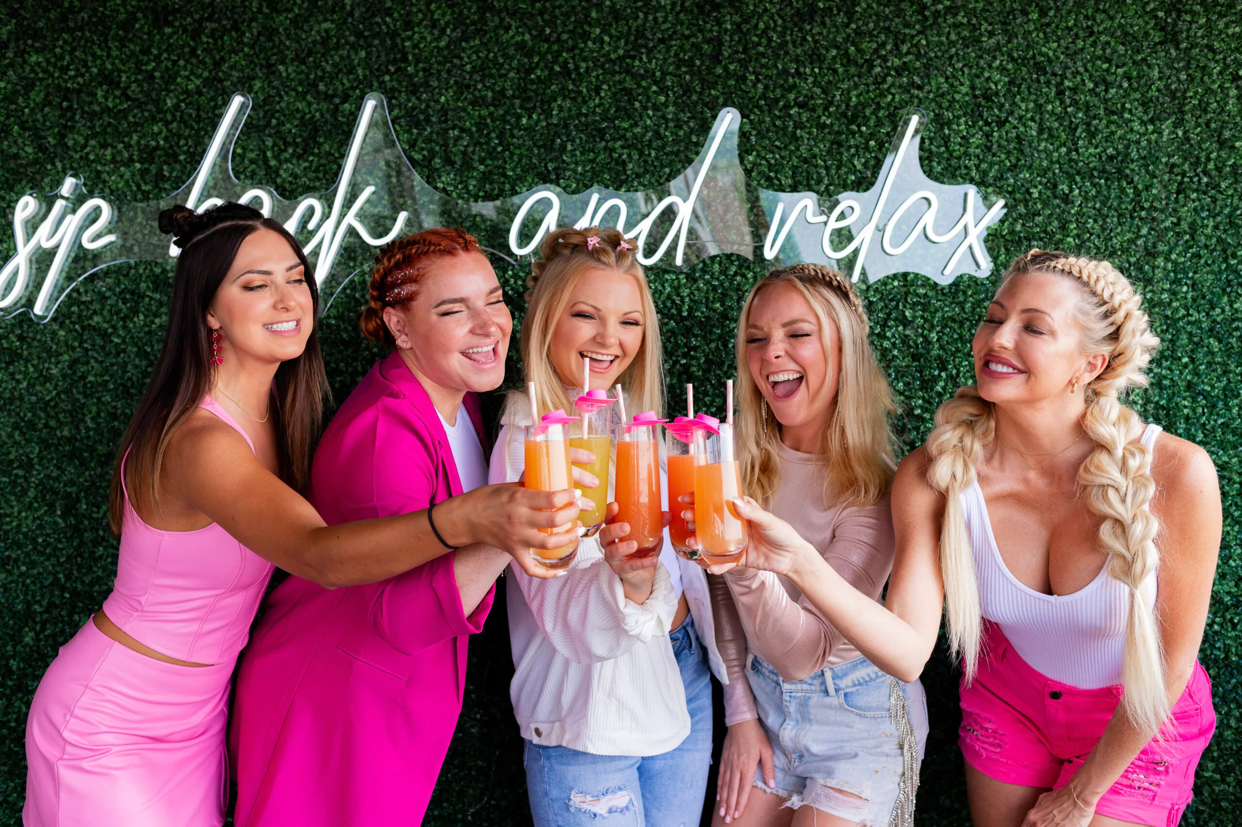Three women in pink outfits toasting with orange drinks in front of a green hedge wall with a "back and relax" neon sign