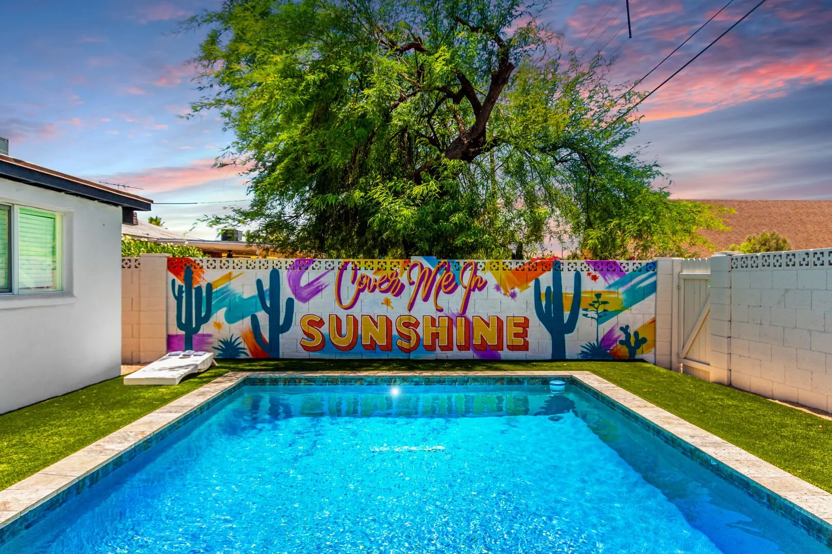 A sunny Palm Springs backyard pool with a colorful "You Are My Sunshine" cactus mural painted on the wall behind it