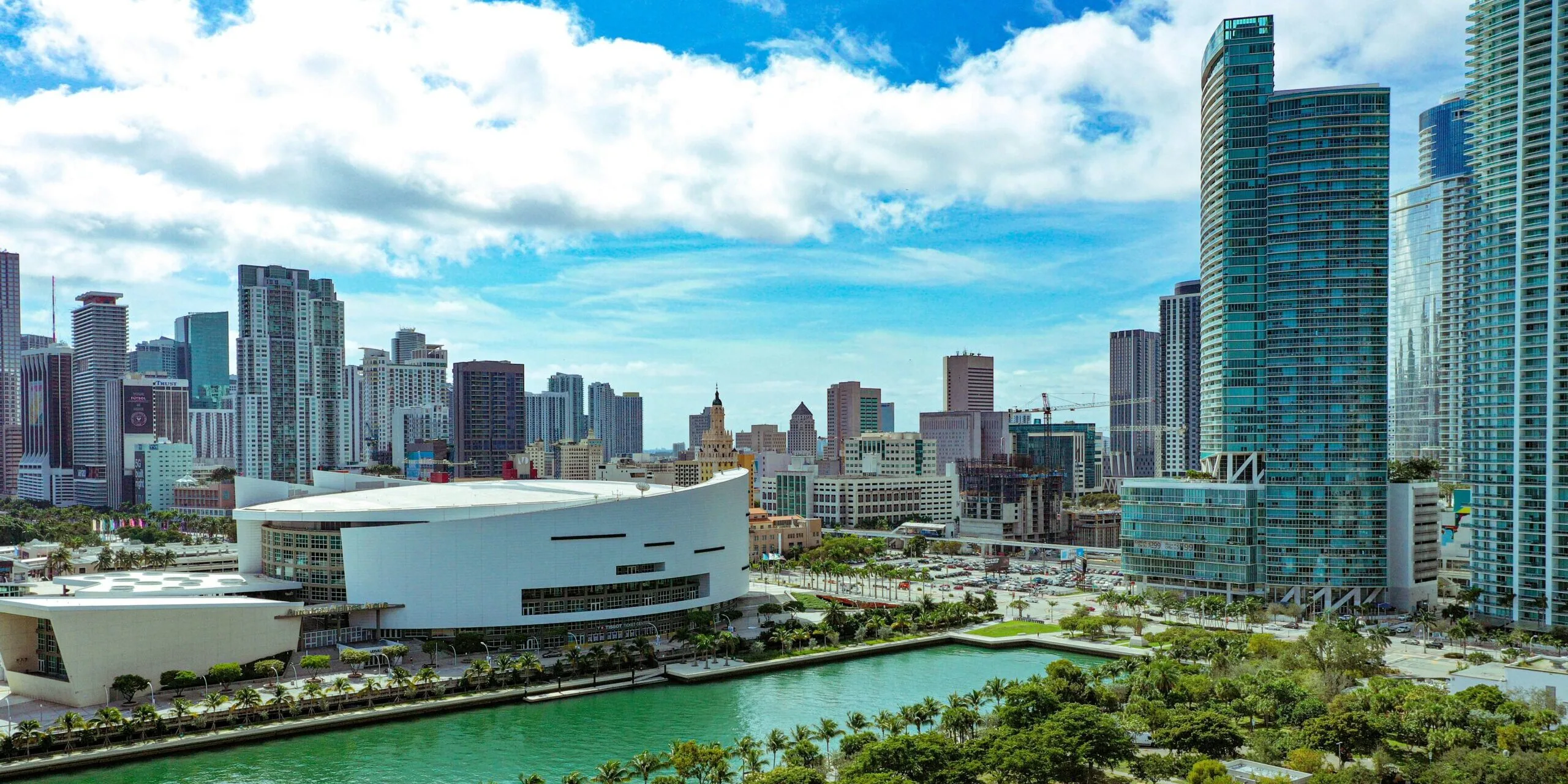 Aerial view of the Miami downtown skyline with the white curved Kaseya Center arena, a waterway lined with palm trees, and gleaming glass high-rise towers under a partly cloudy blue sky.