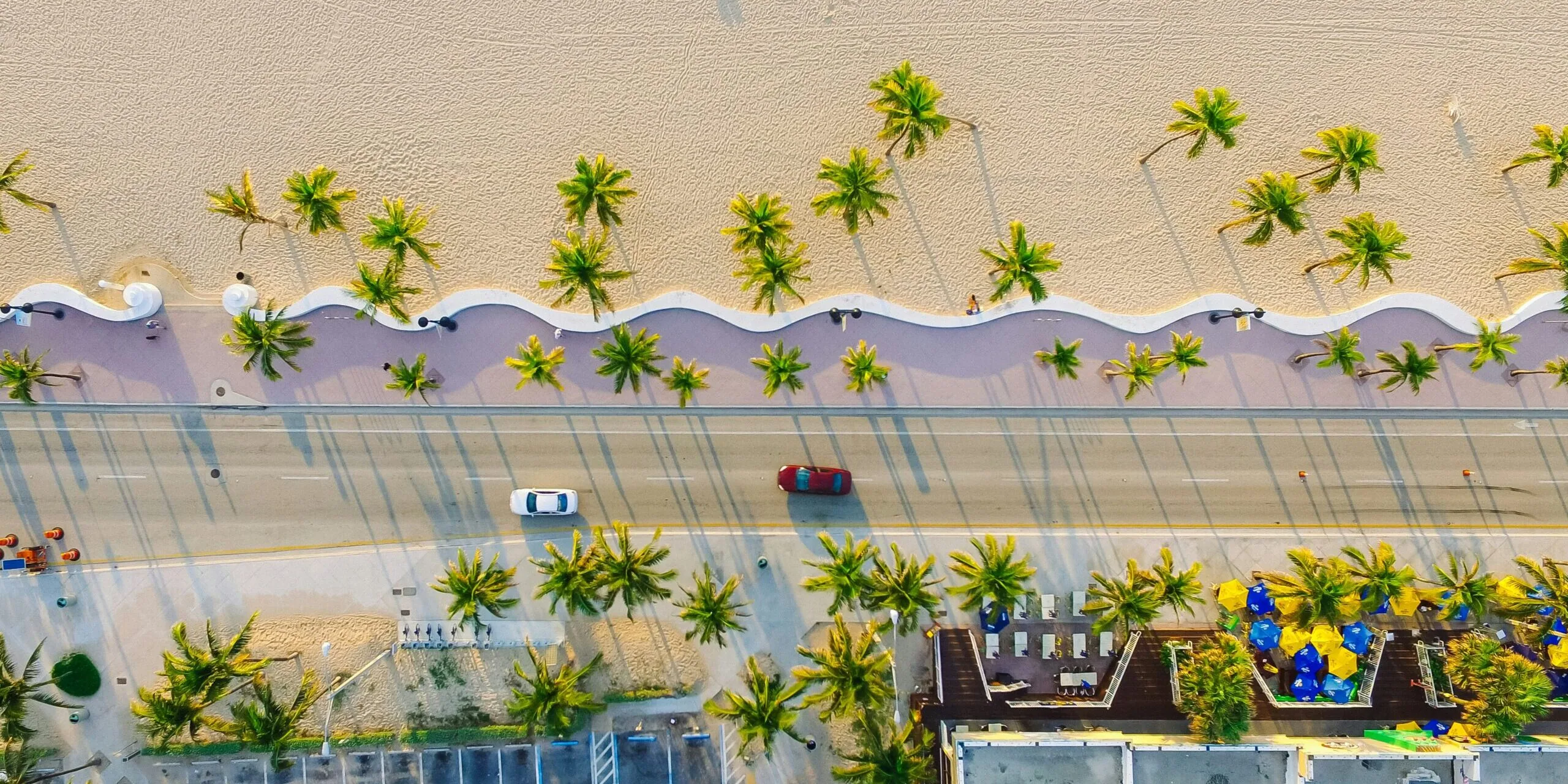 Aerial view of a Miami Beach boulevard with a iconic wave-patterned promenade separating the road from the sandy beach, lined with palm trees and colorful beach umbrellas visible at a beachside venue below.
