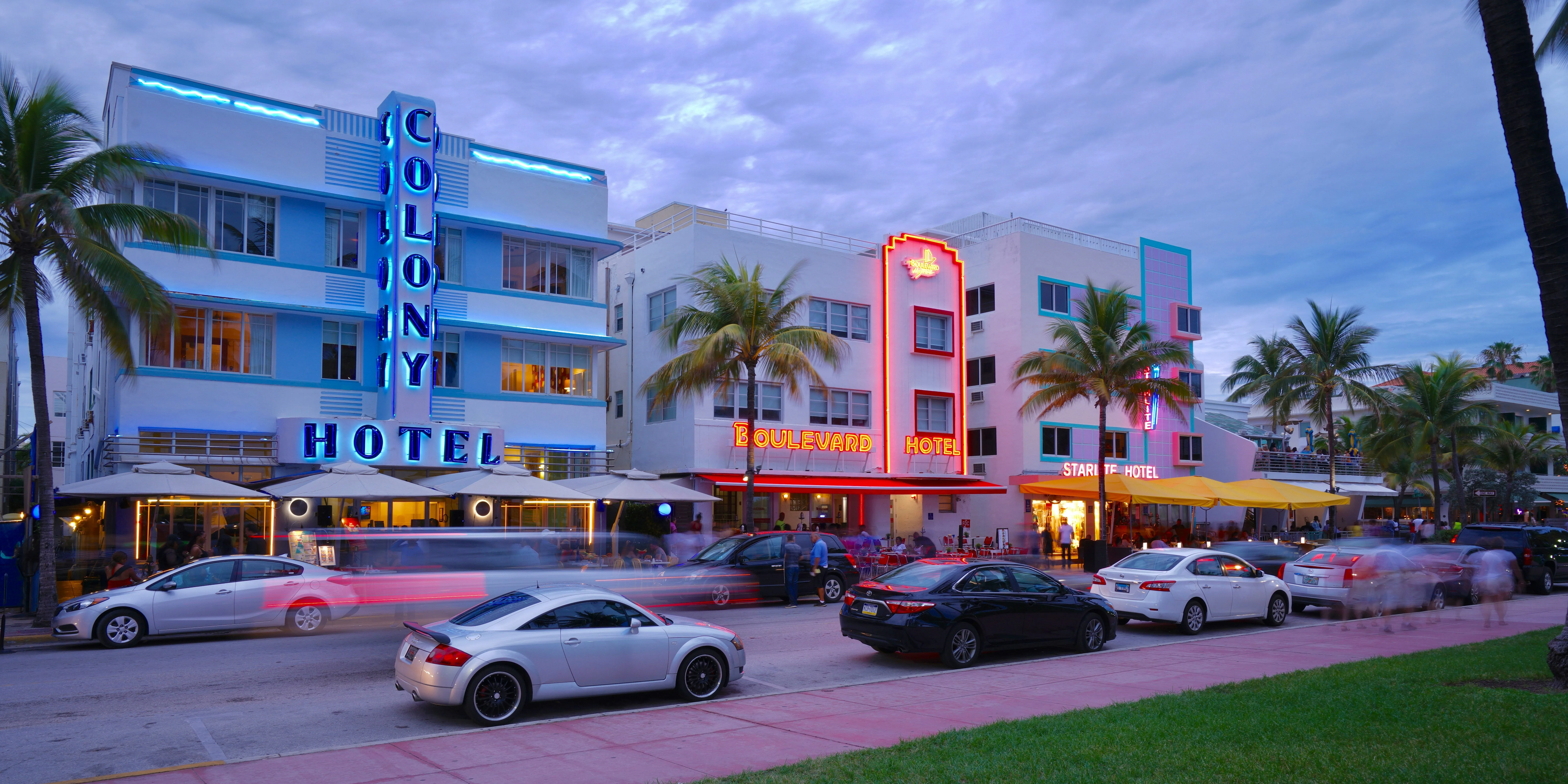 The iconic Art Deco hotels along Ocean Drive in Miami Beach at dusk, including the Colony Hotel and Boulevard Hotel lit up in colorful neon signs, with palm trees and cars lining the street.