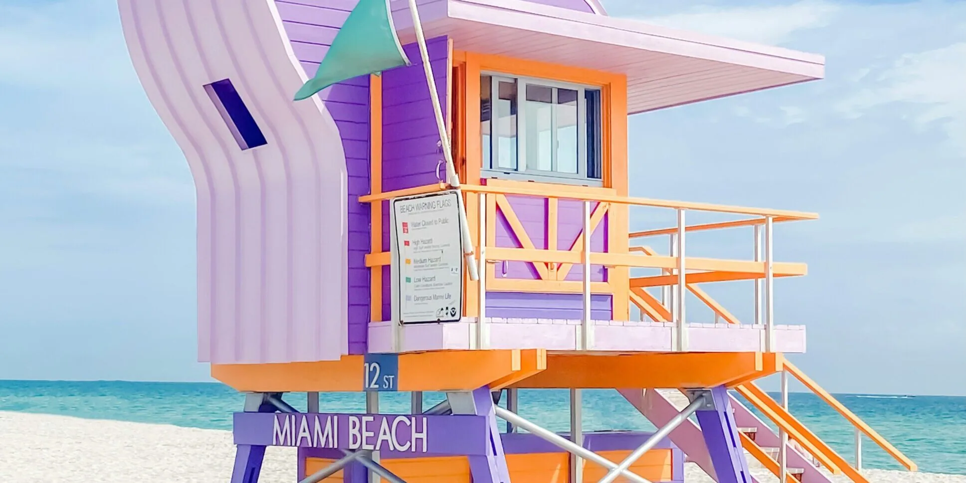 A colorful purple and orange Art Deco lifeguard stand labeled "Miami Beach" at 12th Street, with turquoise ocean water and white sand in the background.