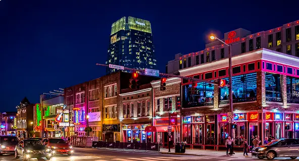 Neon-lit Broadway entertainment district in Nashville at night with glowing bar and honky-tonk signs, a Hilton hotel, and pedestrians on the sidewalk