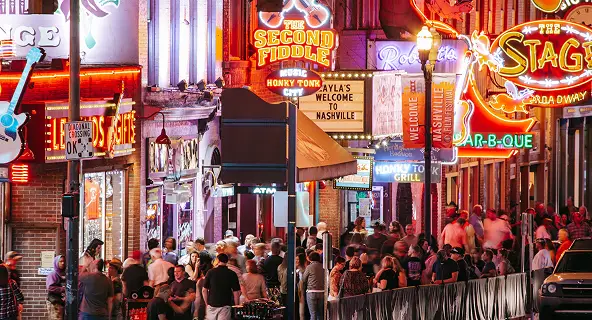 Packed crowd on Nashville's Lower Broadway at night surrounded by glowing neon signs for honky-tonks and live music venues including The Second Fiddle and The Stage
