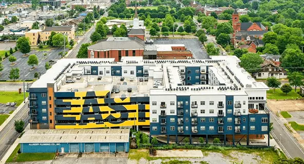 Aerial view of a blue and white mixed-use apartment complex with a bold yellow "EAST" mural on its facade, surrounded by green tree canopy in an urban neighborhood