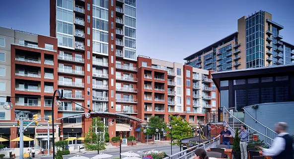 Modern mixed-use urban development with red brick mid-rise apartment buildings, ground-floor retail, and outdoor dining patios in a walkable city neighborhood