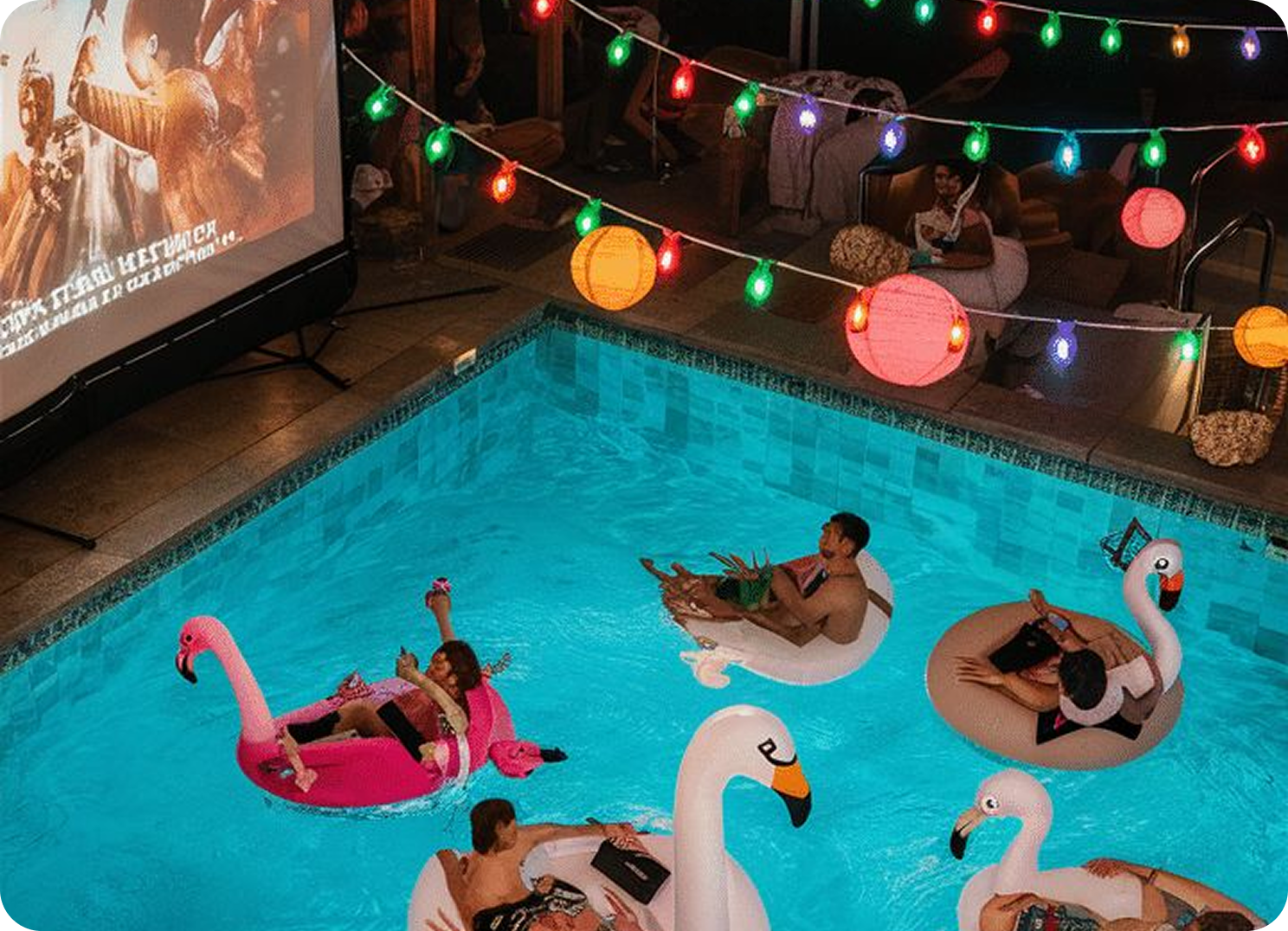 People float on flamingo and swan inflatable pool floats during an outdoor movie night, surrounded by colorful string lights and paper lanterns, with a large projection screen showing a film beside the pool.