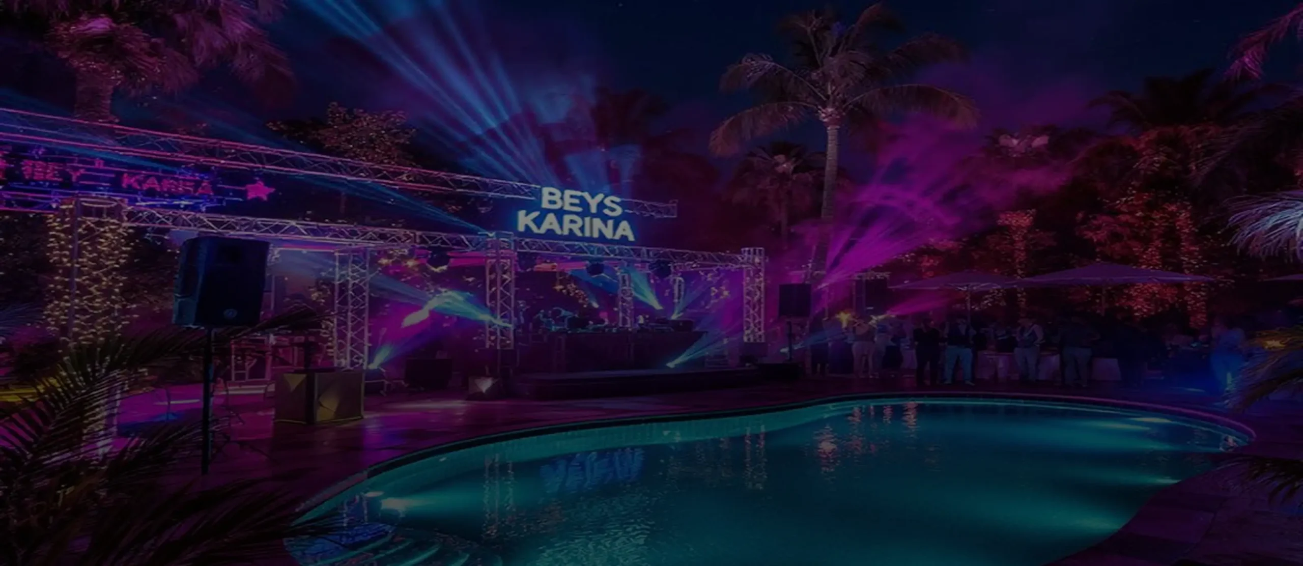 An outdoor nighttime pool party with a DJ stage lit up in purple and blue lights, a custom "Beys Karina" illuminated sign on a truss rig, with palm trees and a glowing pool in the foreground.
