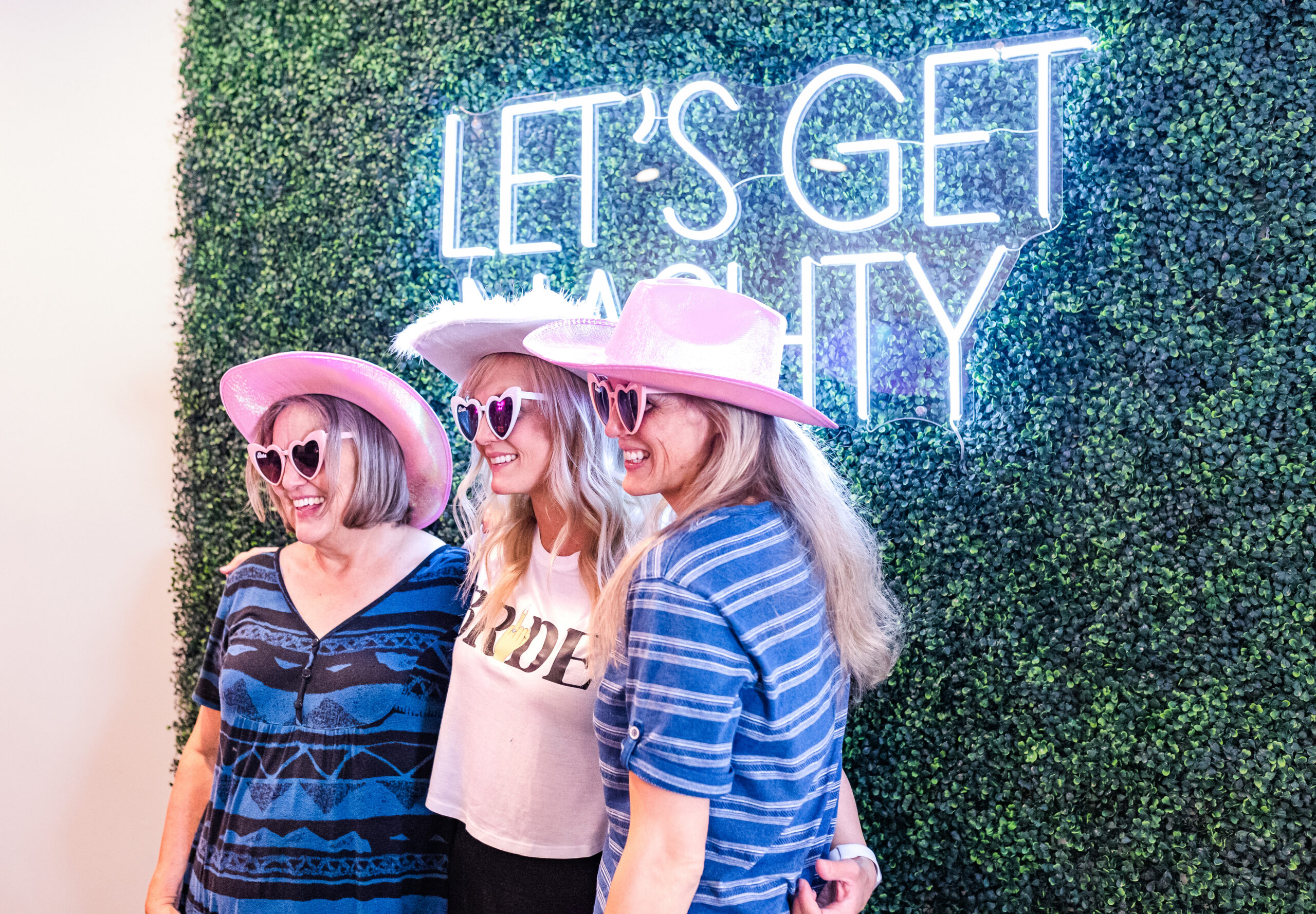 Three women wearing pink cowboy hats pose and smile in front of a neon sign reading "Let's Get Nasty" on a green hedge wall backdrop, with one woman wearing a "Bride" shirt.