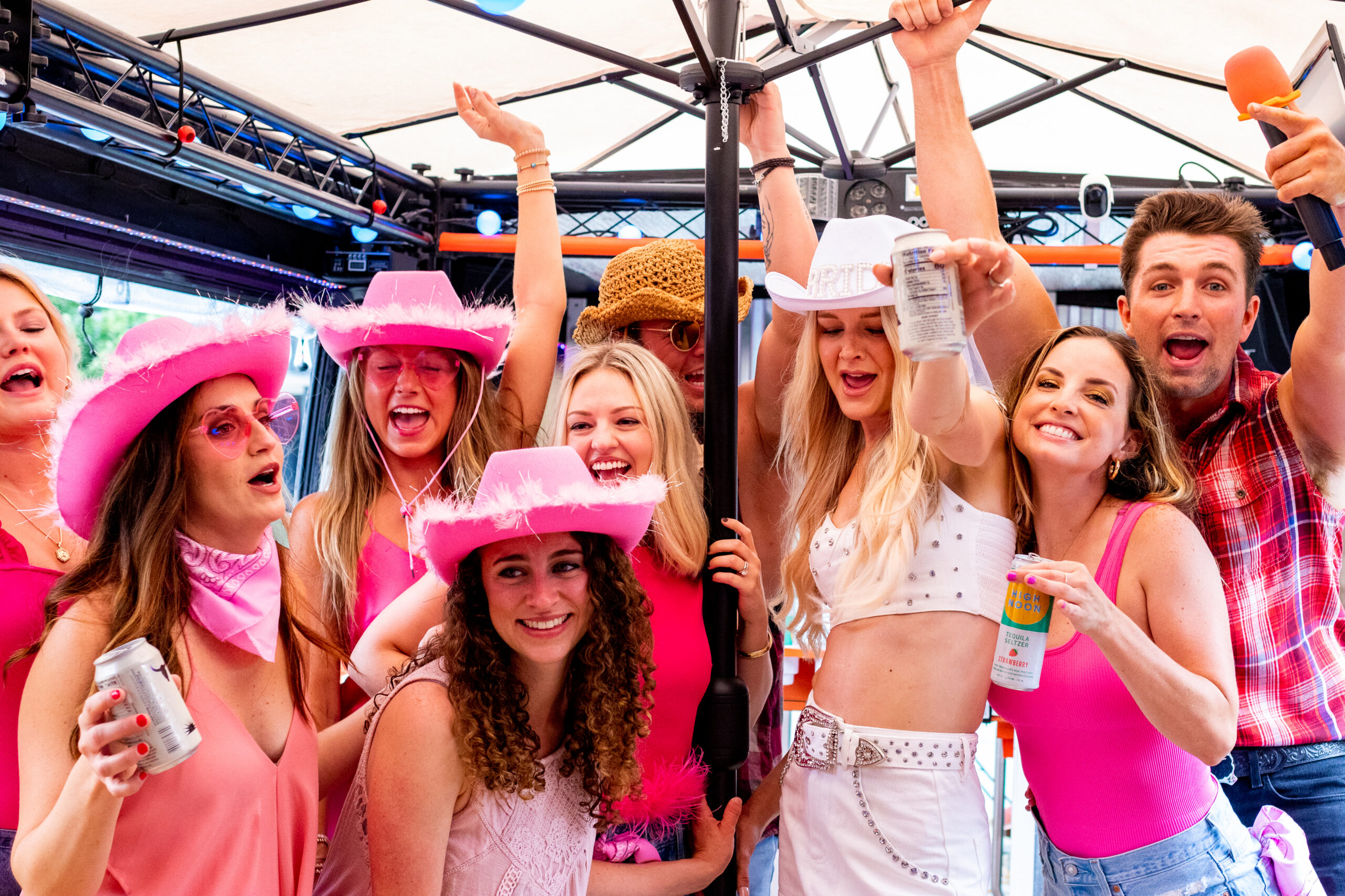 A lively group of partygoers dressed in pink and western-themed outfits celebrating under an open-air tent, laughing and cheering with drinks in hand. Several women are wearing pink cowboy hats, with one guest in a white bedazzled outfit raising her arms in celebration.