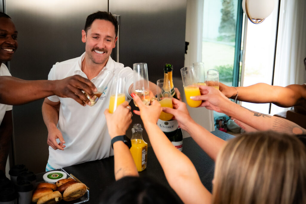 Host in white polo toasts mimosa glasses with guests' hands reaching in around kitchen counter
